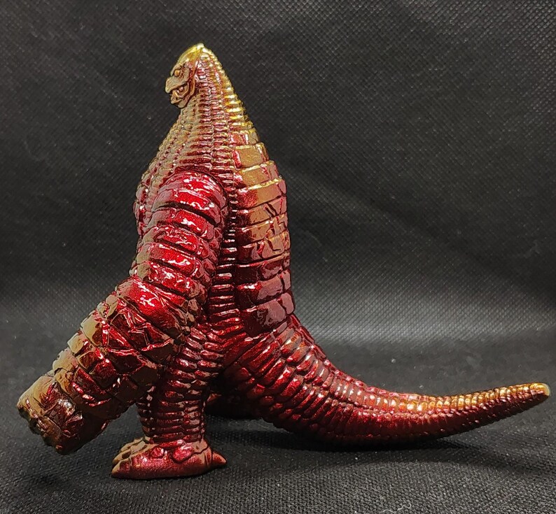 May include: A red and gold figurine of a reptilian creature with a long tail and textured skin. The figure has a detailed head and is posed in a dynamic stance, showcasing its intricate design and metallic finish.