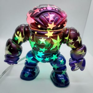 May include: A small, colourful, star-patterned robot toy with a purple, green, and blue iridescent finish. The toy has a light-up feature and is made of plastic.
