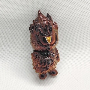 K&ouml;nnte beinhalten: Braune Eulenfigur mit stilisiertem Design. Die Eule hat einen flammenartigen Kopf, ein gro&szlig;es Auge mit goldenem Akzent und einen gefiederten K&ouml;rper. Die Figur steht auf einer wei&szlig;en Oberfl&auml;che.