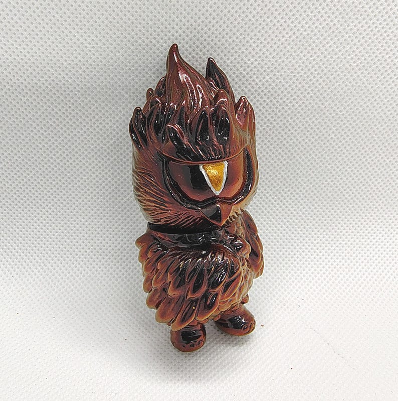 K&ouml;nnte beinhalten: Braune Eulenfigur mit stilisiertem Design. Die Eule hat einen flammenartigen Kopf, ein gro&szlig;es Auge mit goldenem Akzent und einen gefiederten K&ouml;rper. Die Figur steht auf einer wei&szlig;en Oberfl&auml;che.