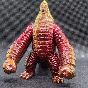 May include: A metallic red and gold monster figurine with a textured, scaly appearance. The figure has a cone-shaped head, thick arms, and stubby legs. The toy is posed in a standing position against a dark background.