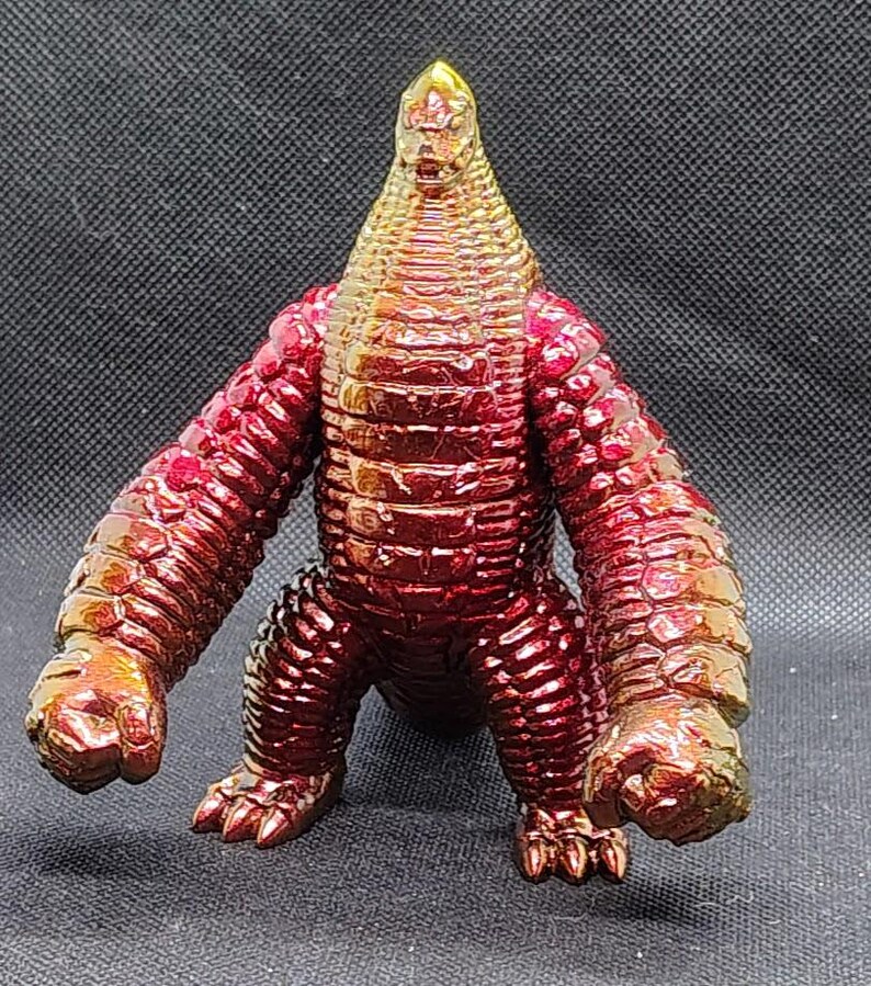 May include: A metallic red and gold monster figurine with a textured, scaly appearance. The figure has a cone-shaped head, thick arms, and stubby legs. The toy is posed in a standing position against a dark background.