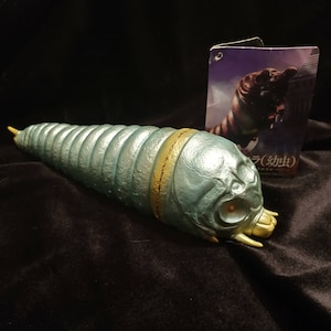 May include: A blue and gold plastic toy of a creature from the Japanese kaiju film Gamera. The toy is posed in a crawling position with its mouth open. The toy is on a black surface. The text on the card behind the toy reads "Gamera (幼虫)".