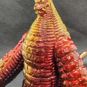 May include: A detailed, close-up of a red and gold monster figurine. The figure has a reptilian head with sharp teeth and a textured body. The color transitions from gold at the head to a deep red on the body and arms.