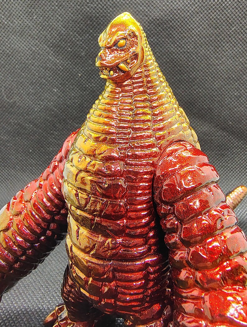 May include: A detailed, close-up of a red and gold monster figurine. The figure has a reptilian head with sharp teeth and a textured body. The color transitions from gold at the head to a deep red on the body and arms.