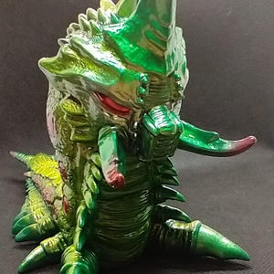 May include: A green and red detailed monster figurine with a long, pointed tail and sharp claws. The monster has a textured, segmented body and a menacing expression.