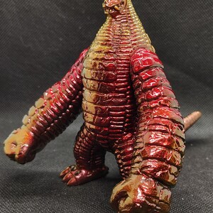 May include: A red and gold monster figurine with a reptilian appearance. The figure has a textured, scaly body, large arms, and a small tail. The head is triangular with a detailed face. The toy is made of a glossy material.