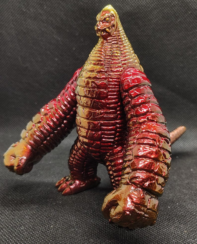 May include: A red and gold monster figurine with a reptilian appearance. The figure has a textured, scaly body, large arms, and a small tail. The head is triangular with a detailed face. The toy is made of a glossy material.