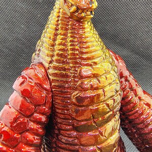 May include: A detailed close-up of a red and gold monster figurine. The figure has a reptilian head with sharp teeth and a textured body. The color transitions from red to gold, creating a striking visual effect.
