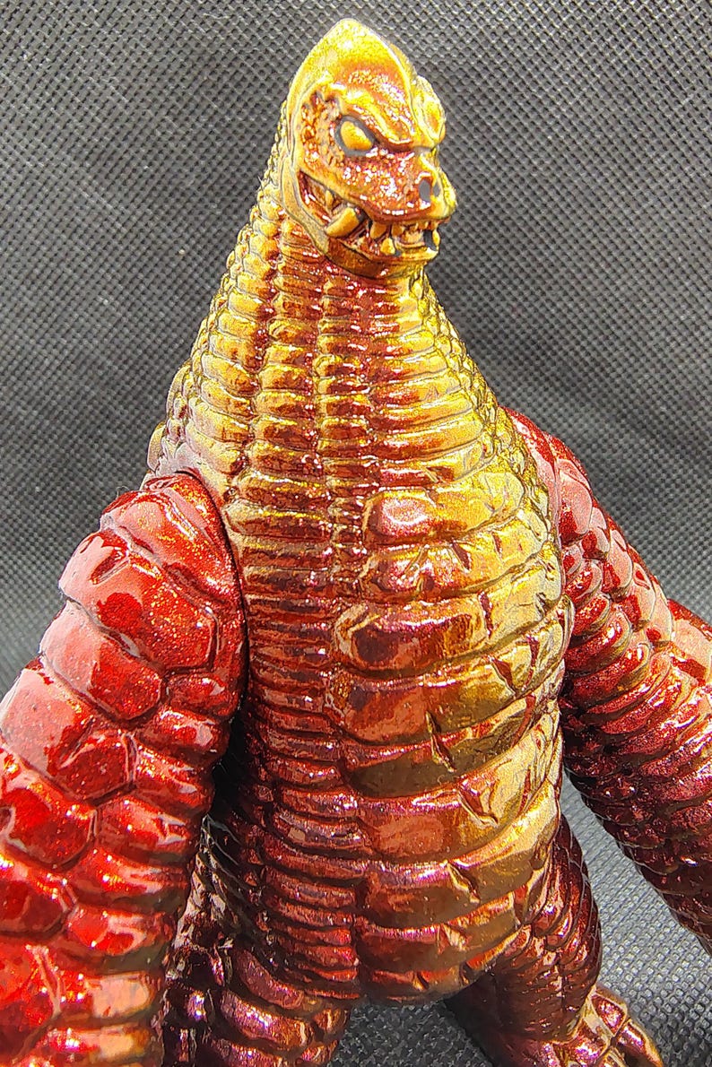 May include: A detailed close-up of a red and gold monster figurine. The figure has a reptilian head with sharp teeth and a textured body. The color transitions from red to gold, creating a striking visual effect.