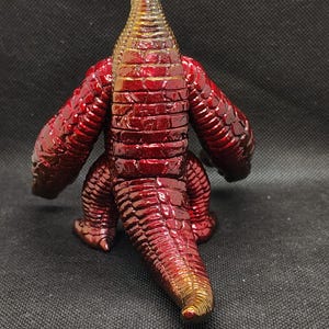 May include: A red and gold figurine of a reptilian creature, viewed from the back. The figure has a textured, scaled appearance with a metallic sheen. The arms and legs are short and stout, and the tail is long. The head is a lighter gold color.