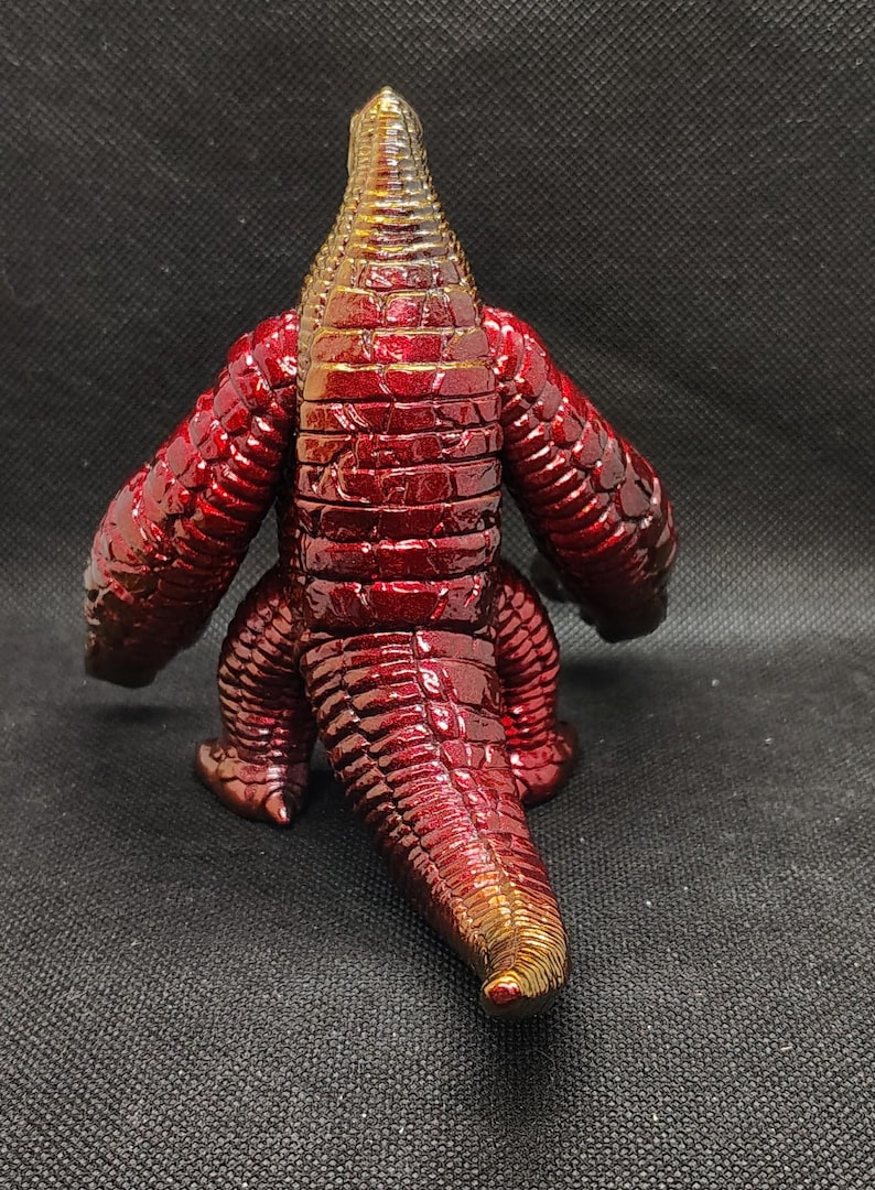 May include: A red and gold figurine of a reptilian creature, viewed from the back. The figure has a textured, scaled appearance with a metallic sheen. The arms and legs are short and stout, and the tail is long. The head is a lighter gold color.