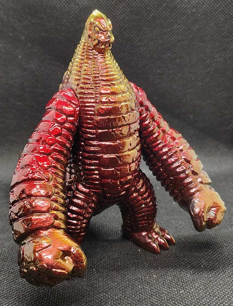 May include: A metallic red and gold monster figurine with a textured, scaly appearance. The figure has a reptilian head, large arms, and a stout body. The toy is posed with its arms at its sides and its feet planted firmly on the ground.