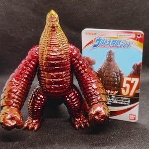 May include: A red and gold action figure of a monster with a textured body, large arms, and a reptilian head. The figure is standing next to a card with Japanese text and the number 57.