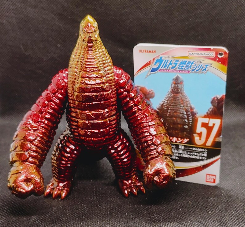May include: A red and gold action figure of a monster with a textured body, large arms, and a reptilian head. The figure is standing next to a card with Japanese text and the number 57.