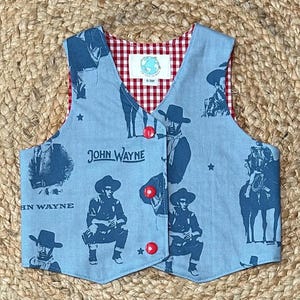 May include: A blue denim vest with a red and white gingham lining. The vest has a button closure and features a repeated pattern of John Wayne images and the text "John Wayne".