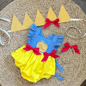 May include: A yellow and blue baby onesie with a red apple and red bows. The onesie is part of a Snow White costume set that also includes a gold crown headband and a red bow headband.