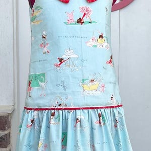 May include: A light blue sleeveless dress with a red peter pan collar. The dress has a ruffled skirt and a whimsical print of children playing with animals and text that says "You are just too much" and "This is a little bit much".