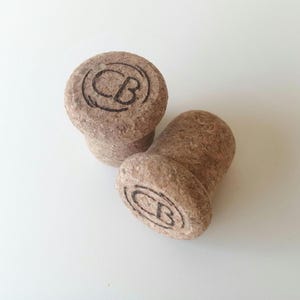 Pode incluir: Dois rolhas de cortiça castanho para garrafas com as letras "CB" gravadas na parte superior.
