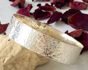 May include: A hammered silver bangle bracelet rests on a light-colored stone, with dried rose petals scattered around. The bracelet has a textured surface and a simple, elegant design, suitable for everyday wear.