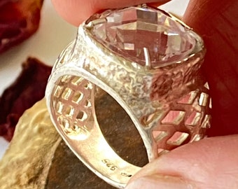 Puede incluir: Un anillo de plata con una gran piedra preciosa facetada transparente. La banda del anillo es texturizada y tiene un diseño recortado. El anillo está marcado con "925" en el interior.