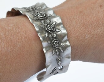 May include: Silver bracelet with a wavy design and a row of raised roses.