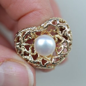 Op de afbeelding: Een gouden ring in de vorm van een hart met een delicaat, getextureerd ontwerp en een witte parel in het midden.