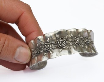 May include: Silver-toned cuff bracelet with a floral rose design. The bracelet has a textured, wavy edge and is decorated with detailed rose and leaf motifs. The open cuff design allows for easy wear.