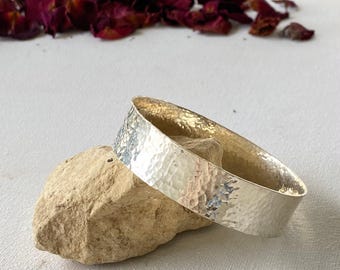 May include: A hammered silver bracelet with a textured surface rests on a light brown stone. The bracelet has a wide band and a shiny finish. Dried rose petals are scattered in the background.