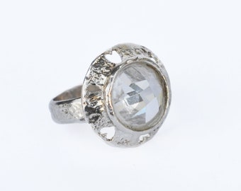 Puede incluir: Un anillo de plata con una piedra preciosa transparente facetada engastada en un engaste circular texturizado. El engaste tiene un diseño decorativo con recortes.