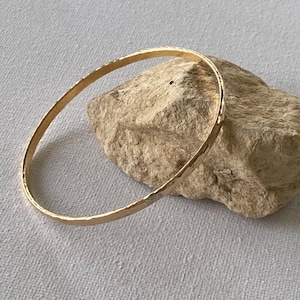 Puede incluir: Un brazalete de color dorado con una textura martillada. El brazalete circular descansa sobre una roca marrón clara sobre un fondo neutro. El brazalete es una pieza de joyería sencilla y elegante.