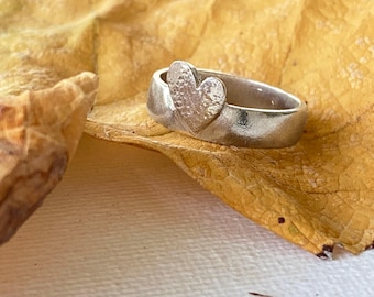 Könnte beinhalten: Ein silberner Ring mit einem strukturierten Herzdesign. Der Ring liegt auf einem gelben Blatt.