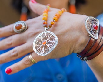 Puede incluir: Un collar de plata con un gran colgante intrincado con un diseño circular con una pequeña piedra preciosa naranja en el centro. El collar está hecho de cuentas naranja y amarillas y se lleva con una pulsera de cuero marrón con un dije de plata.