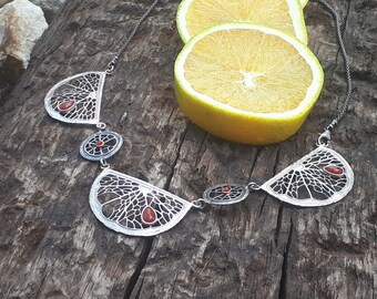 Puede incluir: Un collar de plata con tres colgantes con forma de rodajas de naranja. Cada colgante tiene un delicado diseño de filigrana y una piedra preciosa roja en el centro.