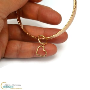 Puede incluir: Un brazalete dorado con textura martillada y un pequeño dije de corazón. El dije de corazón está suspendido de un pequeño anillo sujeto al brazalete. El brazalete se sostiene en una mano sobre un fondo blanco.