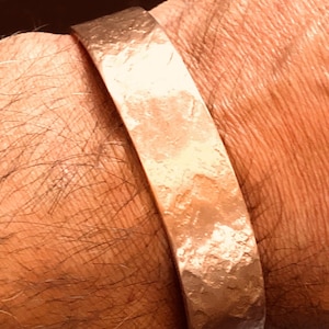 Könnte beinhalten: Ein gehämmertes Kupferarmband. Das Armband hat eine strukturierte Oberfläche und eine warme, rötlich-braune Farbe. Das Armband ist etwa 2,5 cm breit.