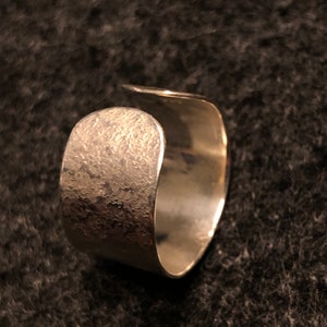Può includere: Un anello d'argento con una finitura testurizzata. L'anello è aperto e ha una forma arrotondata.