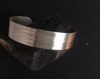 May include: A silver bracelet with a hammered finish. The bracelet is wide and flat, and it has a simple, modern design.