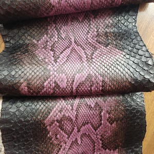 Puede incluir: Primer plano de cuero de piel de serpiente rosa y negro. El cuero tiene un patrón de escamas con una mezcla de tonos rosa, negro y marrón. La textura parece áspera y el material está doblado sobre sí mismo.