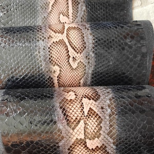Puede incluir: Primer plano de material de cuero enrollado con un patrón de piel de serpiente. El patrón presenta un diseño central beige y marrón flanqueado por escamas de color gris oscuro. El cuero tiene un acabado brillante y los rollos están apilados.
