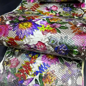 May include: Close-up of a roll of fabric with a snakeskin pattern and a vibrant floral print. The flowers are in shades of purple, pink, yellow, and red, with green leaves. The background is a light gray.