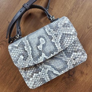 Könnte beinhalten: Eine grau-weiß gemusterte Umhängetasche mit Schlangenmuster und Klappenverschluss. Die Tasche hat einen dunkelgrauen Lederriemen und silberfarbene Beschläge. Die Tasche ist rechteckig und liegt auf einer Holzoberfläche.
