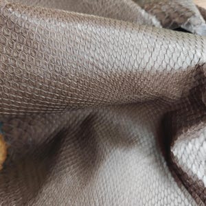 May include: Close-up of brown leather with a snakeskin texture. The leather is folded, revealing the intricate pattern of scales. The colour is a warm, earthy brown, with variations in tone across the surface. The material appears supple and flexible.