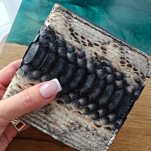 Puede incluir: Una cartera plegable con un patrón de piel de serpiente en tonos grises, negros y crema. La cartera está sujeta por una mano con manicura francesa. El fondo es una superficie de madera con resina epoxi turquesa.