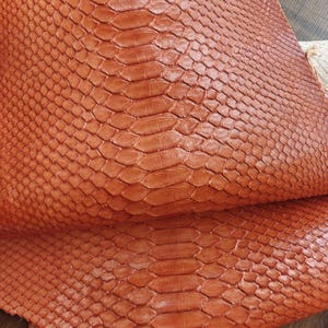 May include: Close-up of a sheet of orange-brown snakeskin leather. The leather has a scaly texture with a repeating pattern of diamond-shaped scales. The scales are slightly raised, giving the leather a three-dimensional appearance.