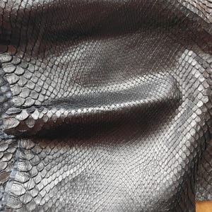 Può includere: Primo piano di pelle di serpente nera e strutturata. Le squame sono visibili, creando un motivo dettagliato. La pelle appare flessibile e presenta leggere variazioni di tono.