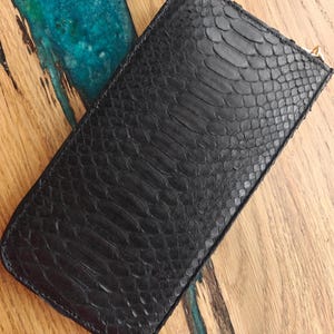 May include: Black leather wallet with a snakeskin pattern. The rectangular wallet has a zipper closure and a gold-colored zipper pull. The wallet is lying on a wooden surface with a blue and green epoxy resin design.