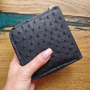 May include: A dark blue, square wallet with a textured surface and a zipper closure. The wallet has a gold-colored zipper pull and is made of a material with a pattern of small, raised dots. The wallet is held in a hand.