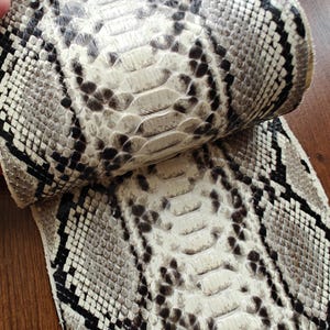 May include: Close-up of a snakeskin pattern. The leather has a natural, textured appearance with a mix of white, black, and grey scales. The scales are arranged in a repeating pattern, creating a unique and eye-catching design.