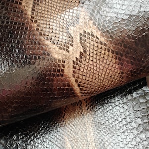 Puede incluir: Primer plano de un material de imitación de piel de serpiente texturizado. La superficie tiene un patrón de escamas en tonos de negro, marrón y beige. El material parece estar doblado, revelando la parte inferior y agregando profundidad a la imagen.